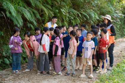 Grupo de crianças participa de uma atividade educativa em uma trilha natural, acompanhado por dois adultos.