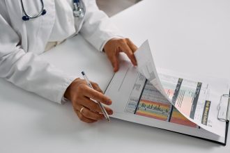 Profissional de saúde verifica relatórios médicos em uma prancheta, em ambiente de atendimento.