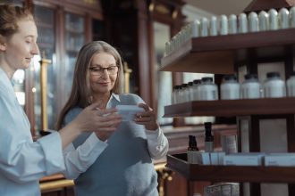 Mulher idosa recebe orientação de um farmacêutico em uma farmácia com prateleiras de medicamentos.