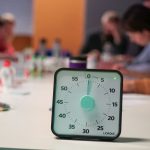 Cronômetro sobre uma mesa, com pessoas desfocadas reunidas em uma sala de escritório ao fundo.