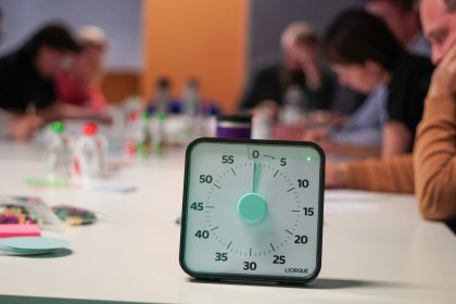 Cronômetro sobre uma mesa, com pessoas desfocadas reunidas em uma sala de escritório ao fundo.