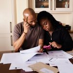 Casal consulta documentos e usa um celular enquanto organiza as finanças domésticas à mesa.
