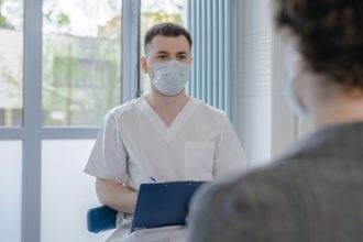 Profissional de saúde de avental consulta uma paciente e faz anotações durante o atendimento.