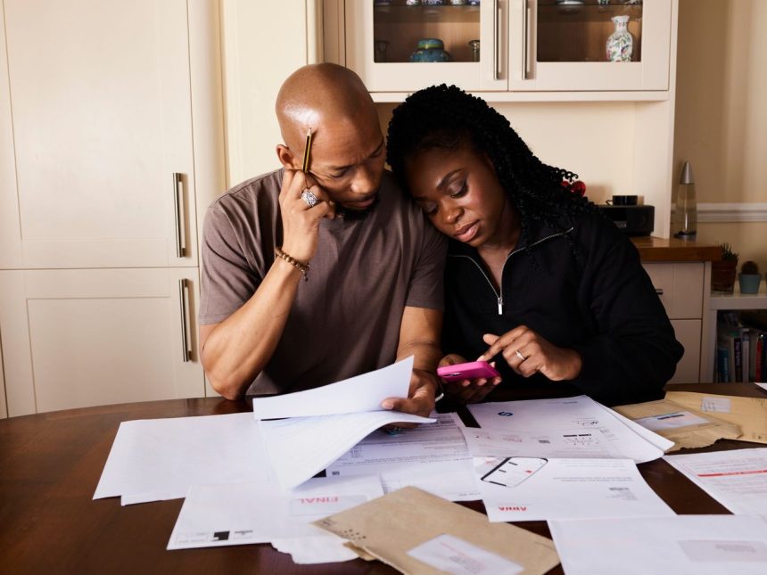 Casal consulta documentos e usa um celular enquanto organiza as finanças domésticas à mesa.