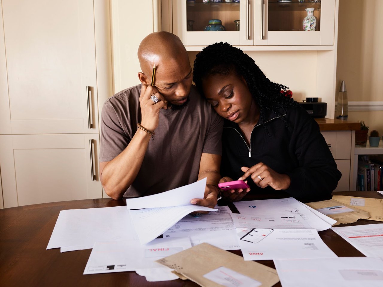 Casal consulta documentos e usa um celular enquanto organiza as finanças domésticas à mesa.